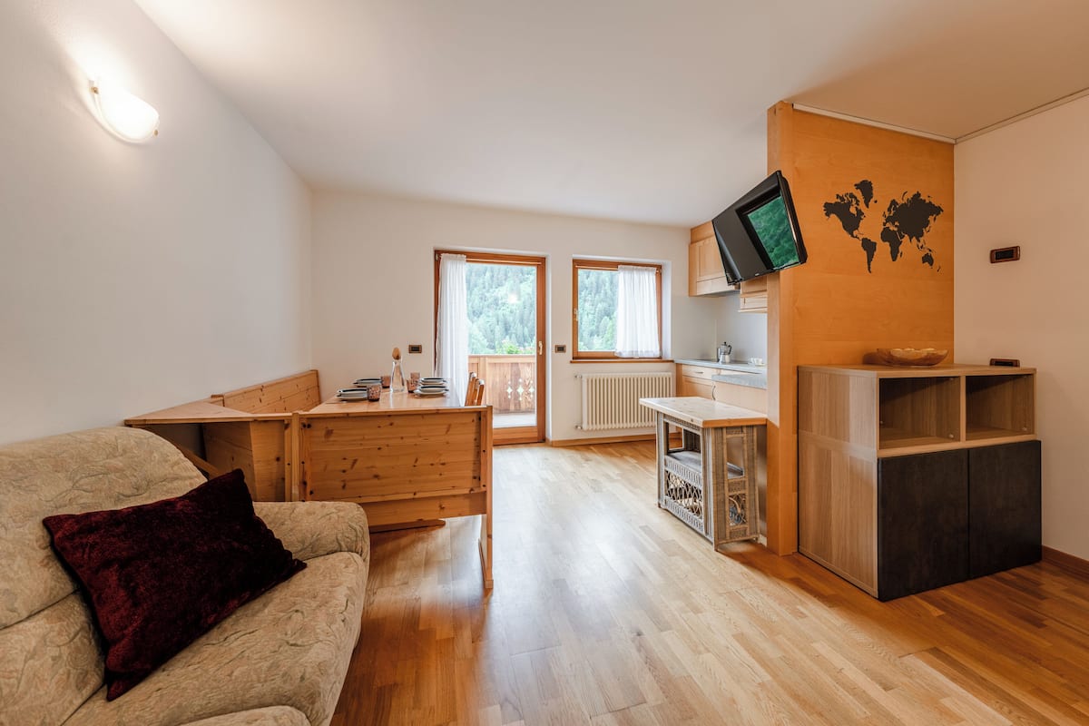 Een knus woongedeelte met een houten vloer, een bank, een eettafel, een keukenhoek met een televisie, en een wereldkaart aan de muur.