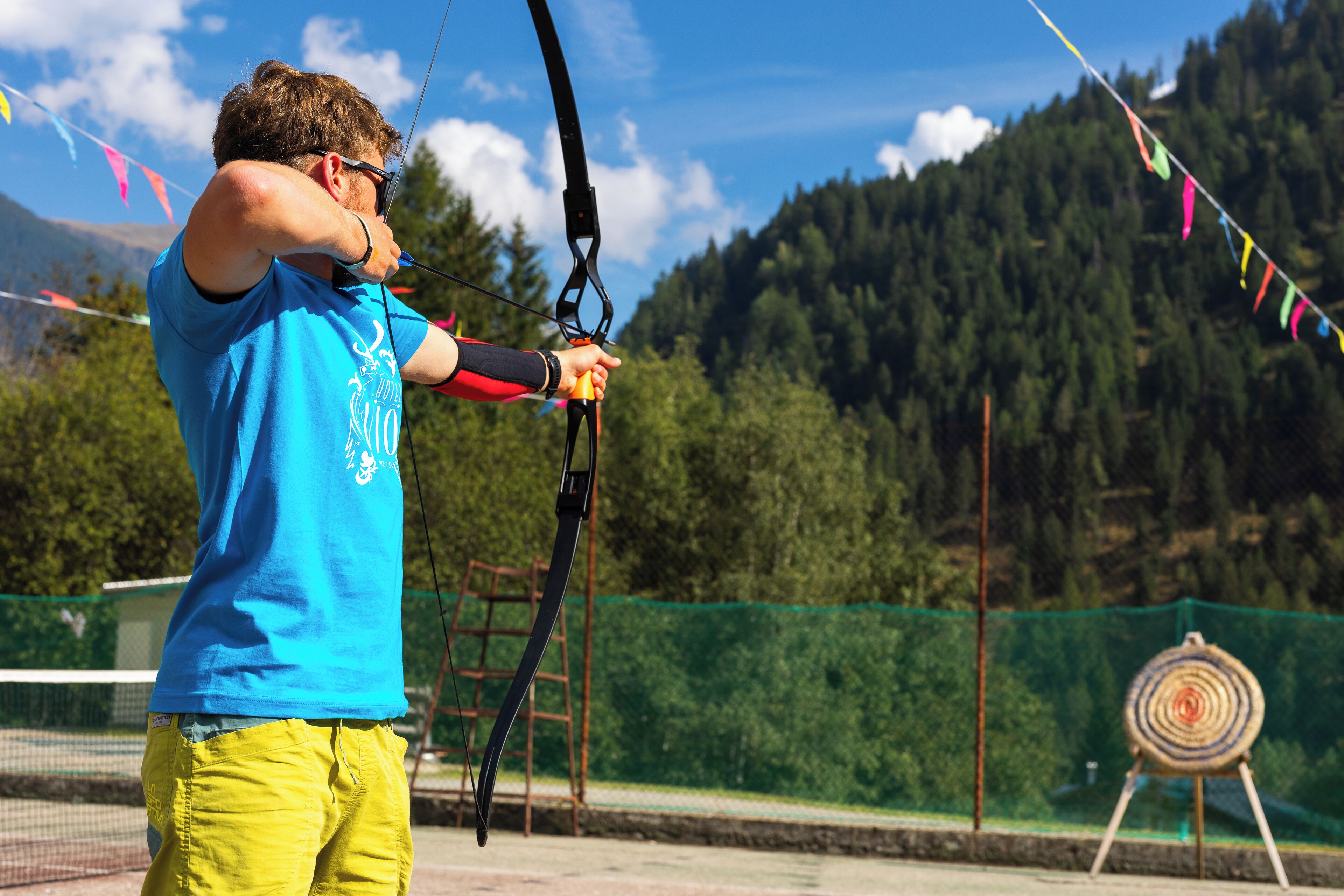 A person in a blue shirt and yellow shorts aiming a bow at a target.