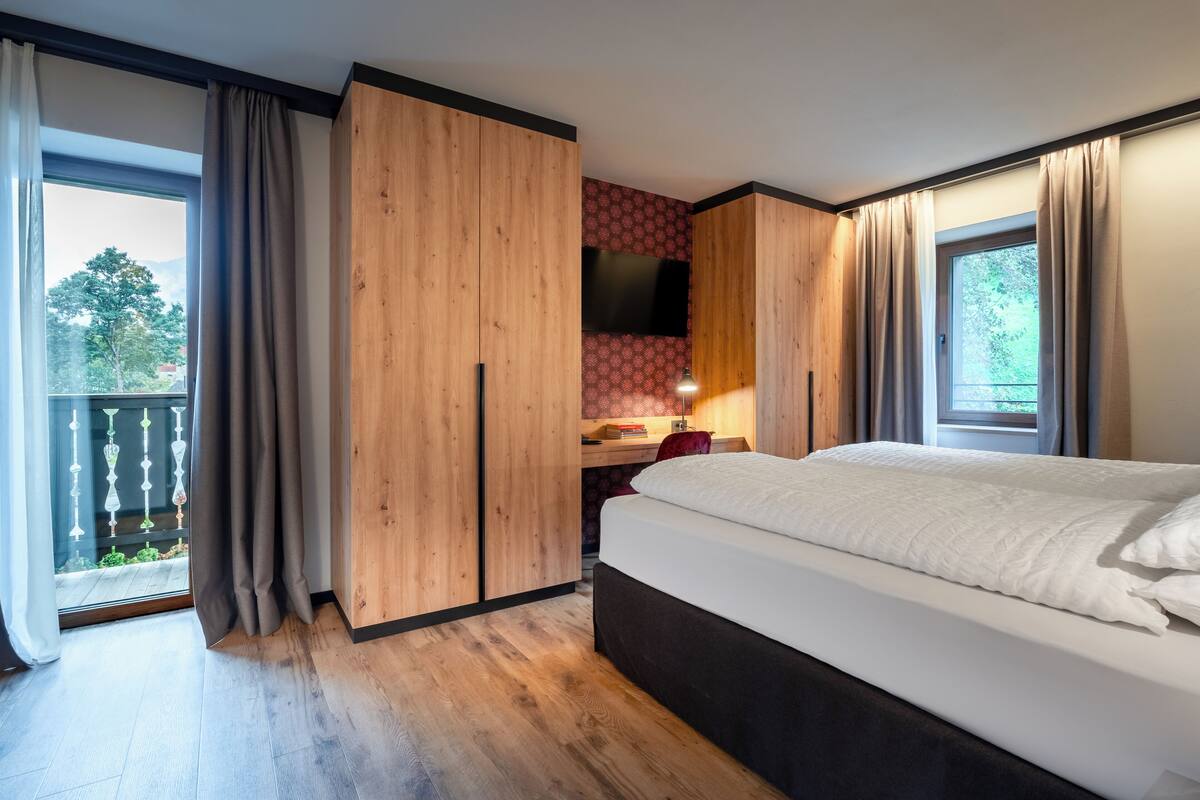 Een slaapkamer met een bed, een houten garderobe, een bureau met stoel, een televisie en een balkon met gordijnen.