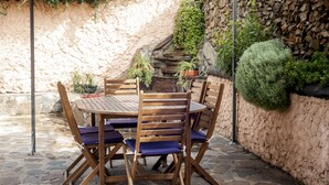 Et sæt træborde og stole på en stenbelagt terrasse med en rustik stenvæg og grønne planter.