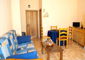 A living room with a sofa, a table, a dining table with chairs, a TV, and a wooden door. - Apartamentos Rurales Azabal (Casar de Palomero)