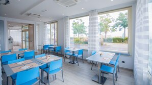 Un espace de restauration moderne avec des chaises bleues et des tables blanches, de grandes fenêtres ornées de rideaux légers, et une vue sur la verdure à l’extérieur.