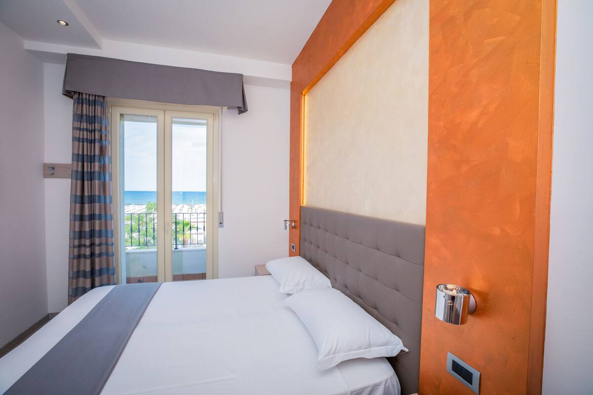 A hotel room with a bed, a nightstand, a wall-mounted light, and a view of the sea through a door.