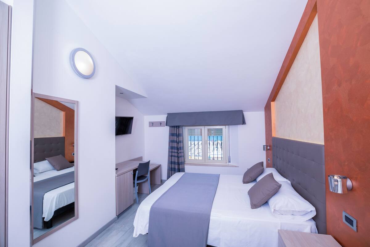 A modern hotel room with a large bed, a desk, a chair, a TV, and a window with curtains.