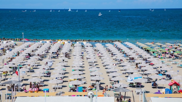 Une plage bordée de rangées de parasols et de chaises longues, un ciel bleu azur et des voiliers sur l’eau.