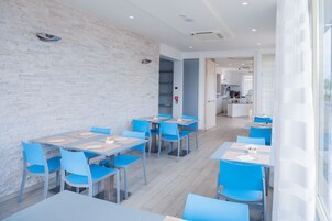 A modern dining area with light-colored brick walls, blue chairs, and tables. There is a kitchen visible in the background.