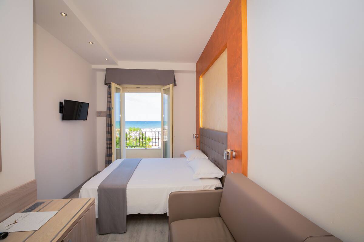 A hotel room with a bed, a desk, a TV, and a view of the beach.