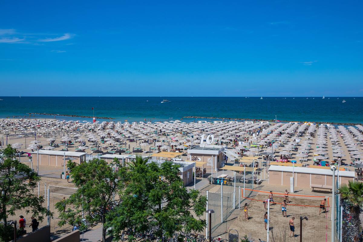 A beach with numerous umbrellas and sun loungers, a volleyball net, and a clear view of the sea.