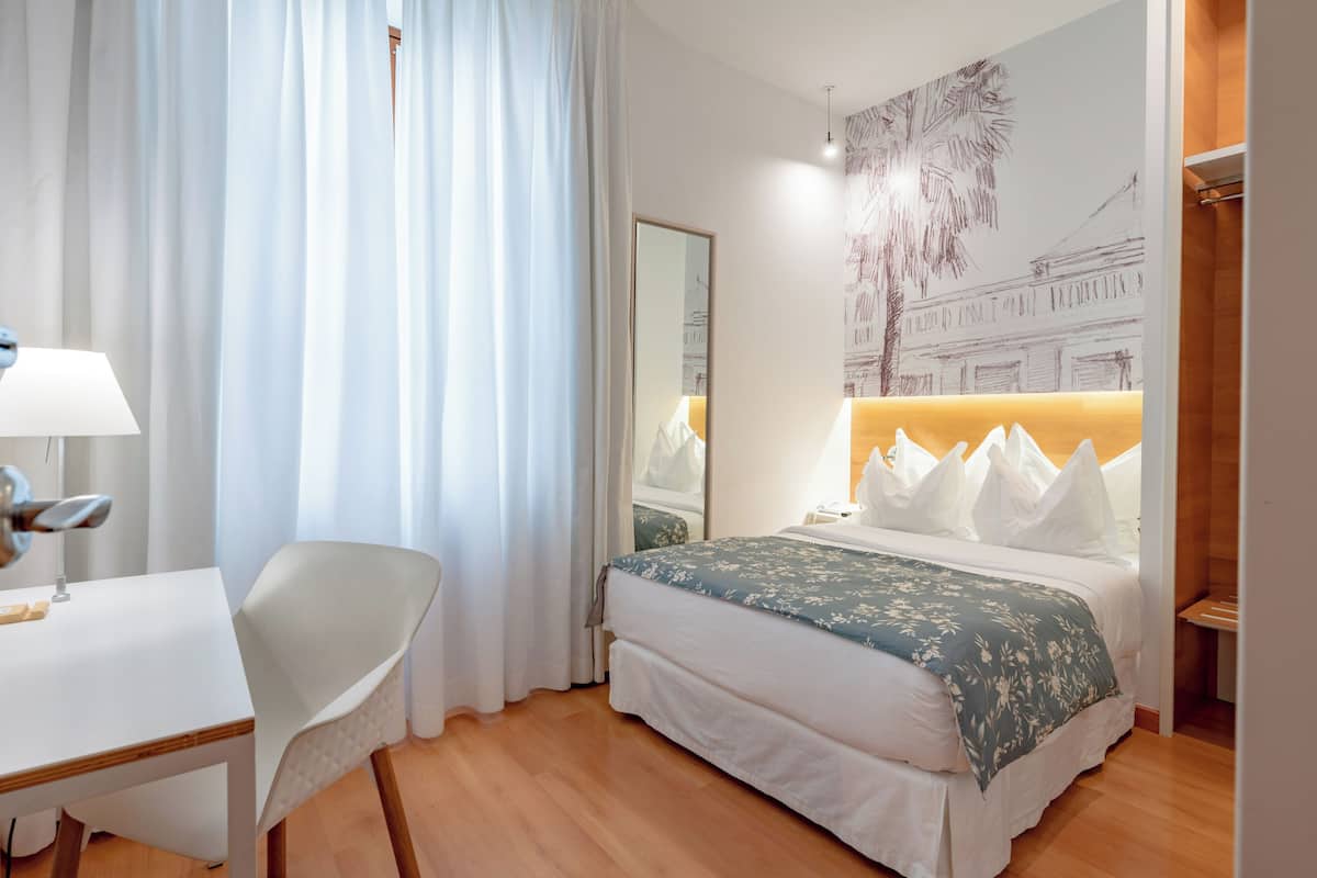 A hotel room with a bed, a desk, a chair, and a large window with curtains.