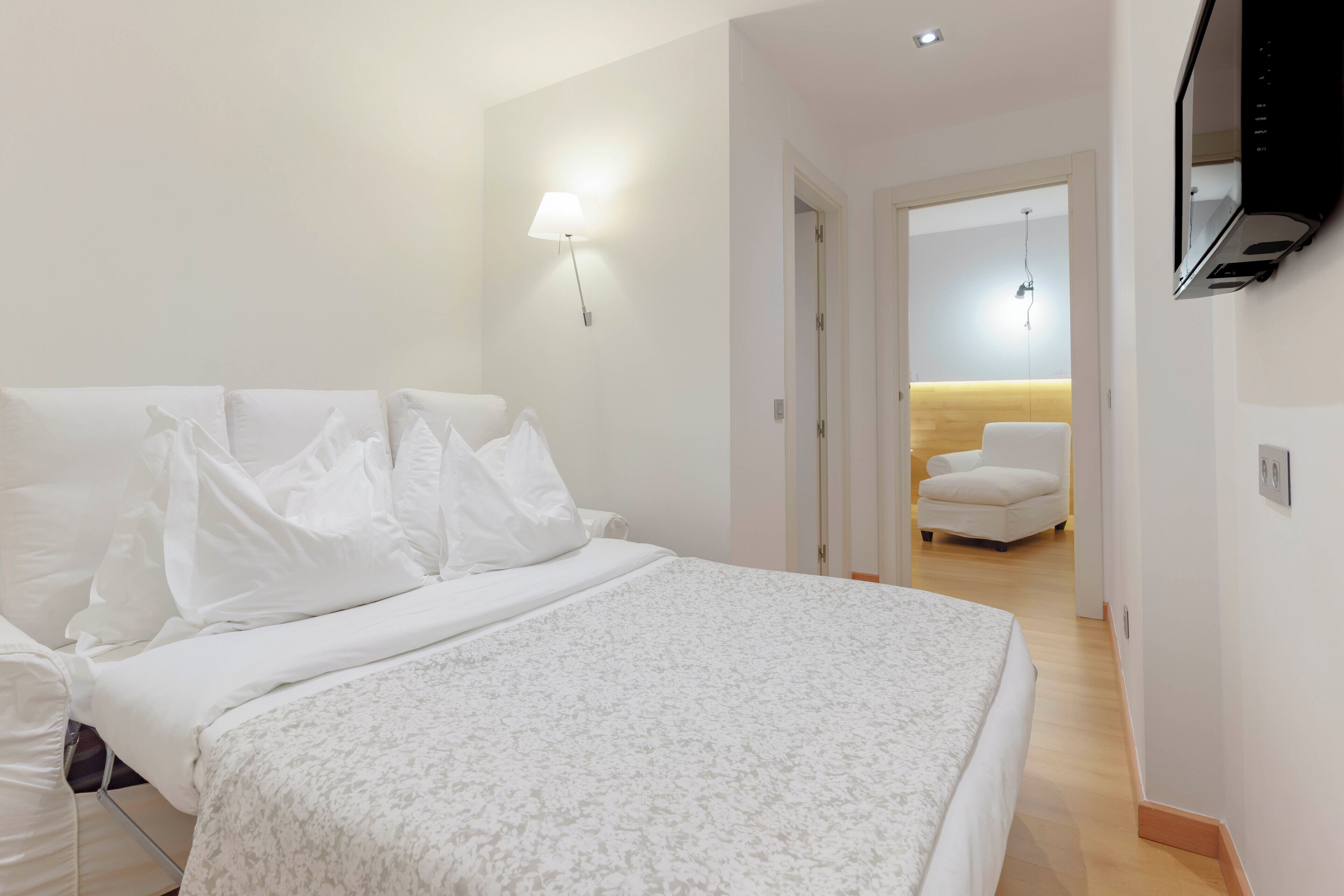 A hotel room with a large bed, a TV mounted on the wall, a chair, and a desk with a lamp.
