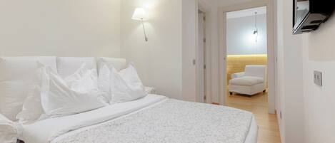 A hotel room with a large bed, a TV mounted on the wall, a chair, and a desk with a lamp.
