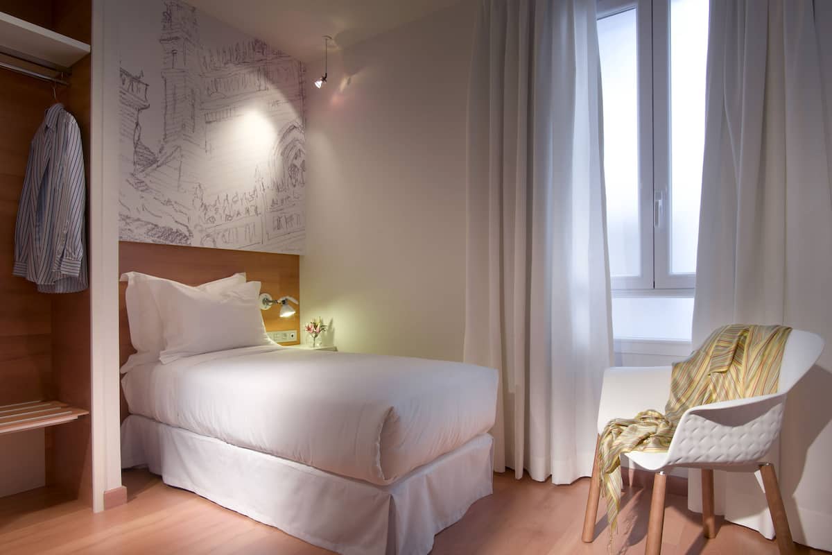 A modern bedroom with a large bed, a bedside table with a lamp, a chair, and a window with curtains.