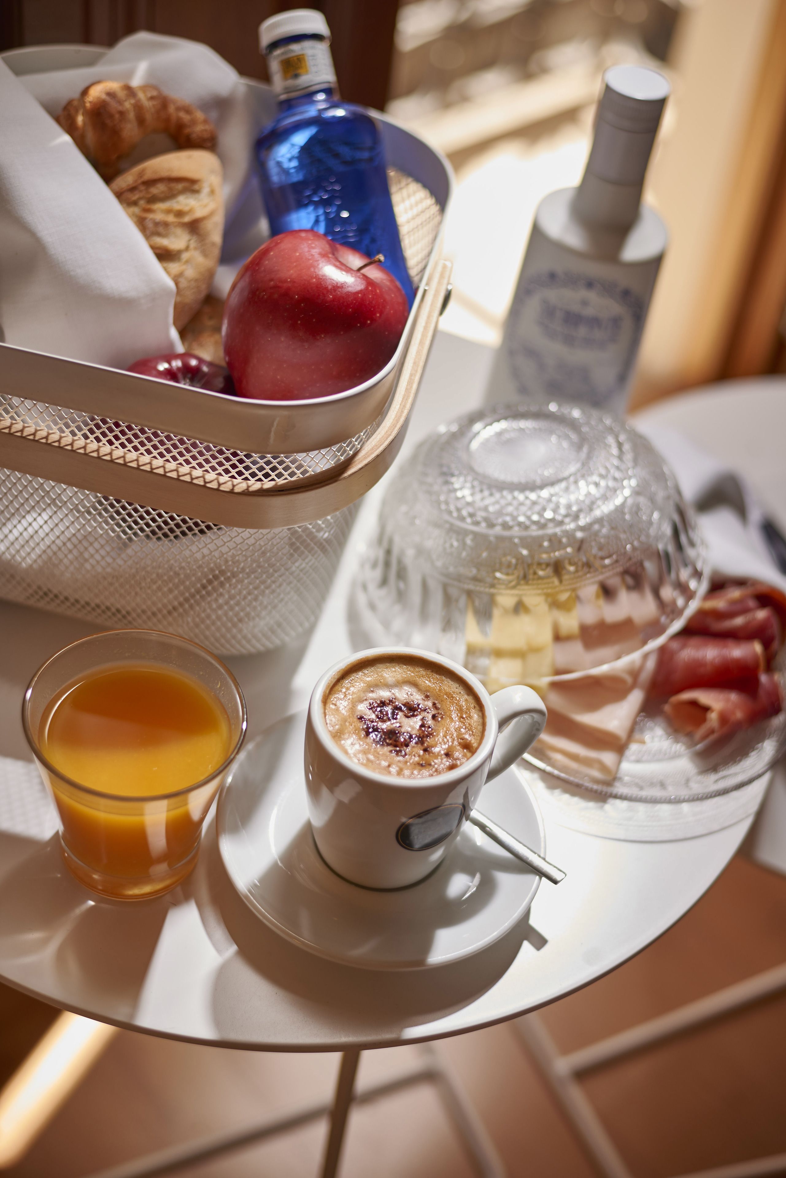 Un vassoio colazione con croissant, una mela, una bottiglia d'acqua, una bottiglia d'olio d'oliva, un bicchiere di succo d'arancia e una tazza di caffè su un tavolo.
