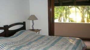 A bedroom with a bed, a nightstand, a lamp, and a view of palm trees through a window. - Pousada do Golfista (Guaruja)