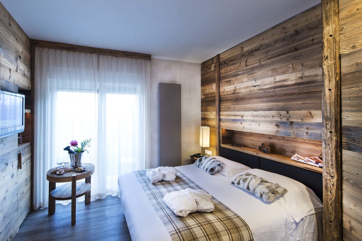Une chambre d’hôtel avec un mur en bois, un lit recouvert de draps blancs, une petite table ronde ornée d’un vase de fleurs et un téléviseur fixé au mur.