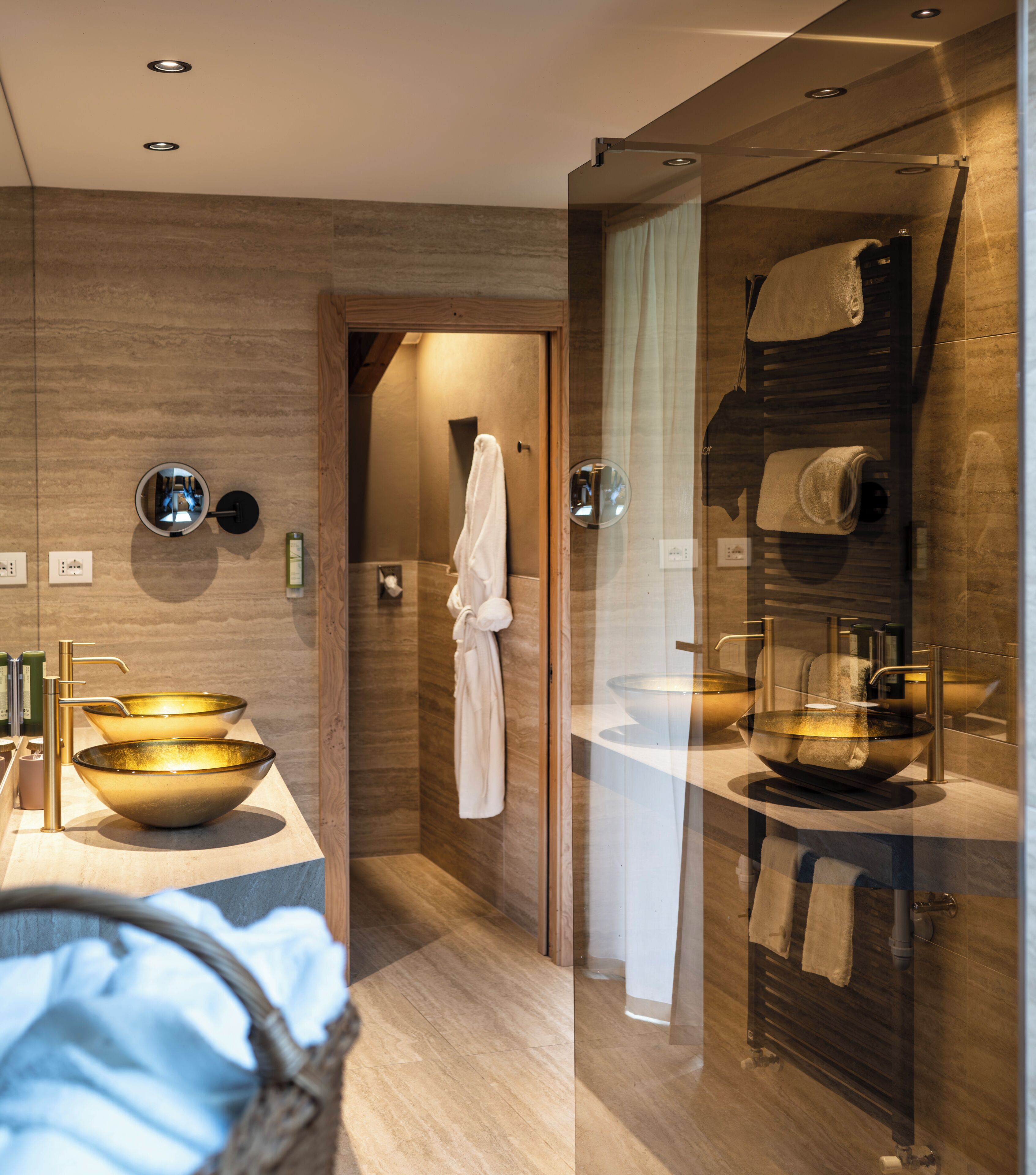 A modern bathroom with a glass shower enclosure, a white robe hanging on a rack, and two large gold sinks.