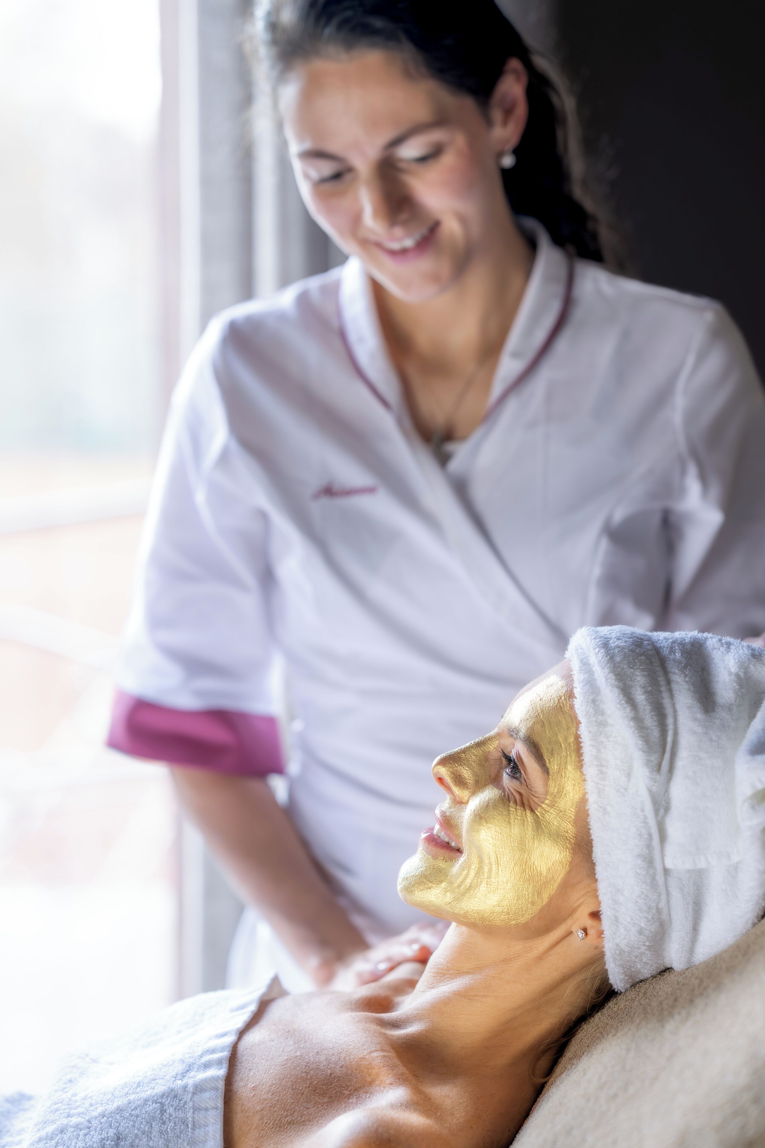 A person receiving a facial treatment with a gold mask, lying on a bed in a room with a window.