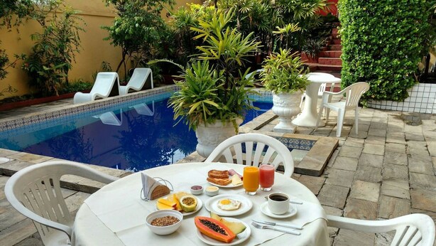 Une table ronde recouverte d’une nappe blanche, entourée de chaises en plastique blanches, est installée au bord de la piscine. Un buffet de petit-déjeuner y est dressé, avec des toasts, des fruits, des œufs et diverses boissons.