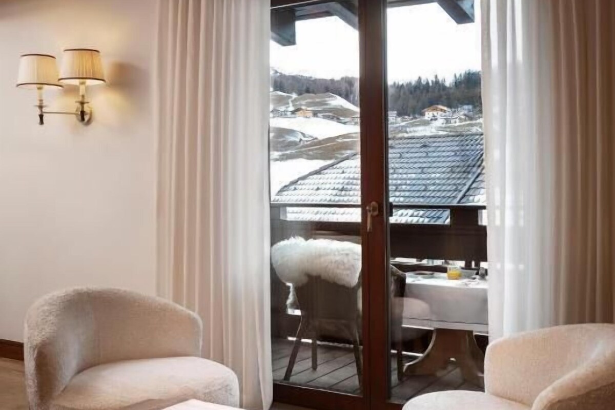 Una habitación con techo de madera, una mesa de comedor con sillas, un cuenco de fruta y vista a un paisaje nevado a través de una puerta de vidrio.