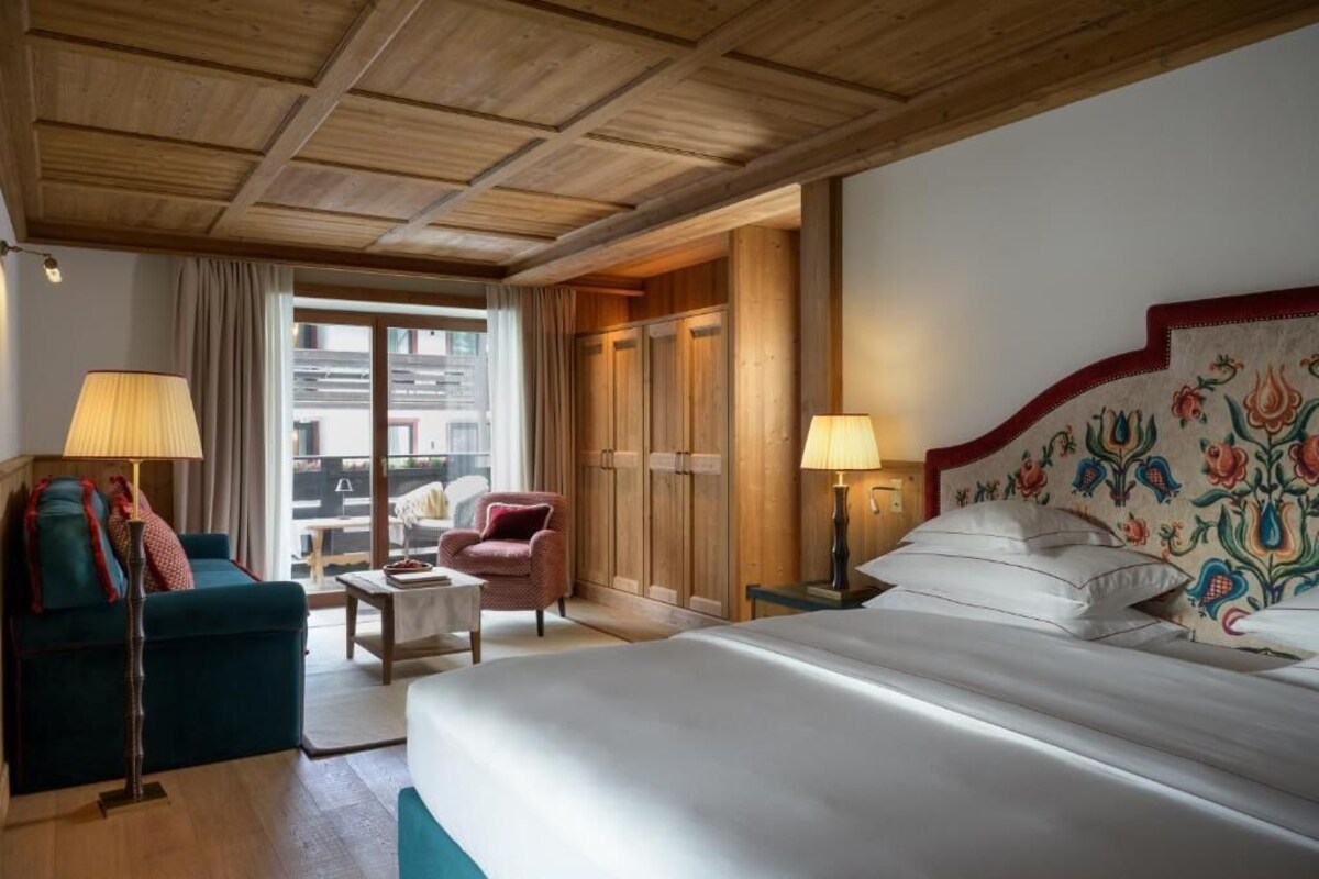 Habitación de hotel con cama, sofá, sillón y escritorio. Cuenta con una ventana con cortinas, techo de madera y una pared con estampado floral.