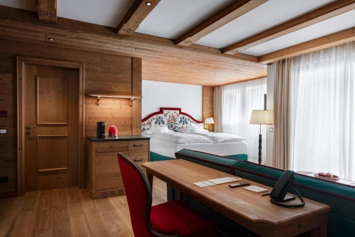 Habitación de hotel con techo de madera, cama con cabecera floral, escritorio con silla y ventana con cortinas.