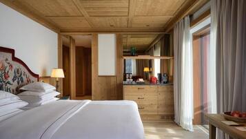 Ein Hotelzimmer mit Holzboden, einem Bett mit weißer Bettwäsche, einem Nachttisch mit Lampe, einem kleinen Tisch mit Äpfeln und Blick ins Freie durch große Fenster.