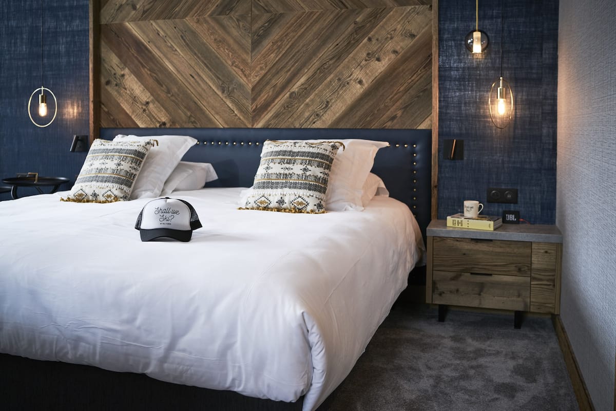 A neatly made bed with white linens and two patterned pillows, a wooden headboard, a small wooden nightstand with a cup and a book, and a wall with a herringbone pattern and hanging lights.