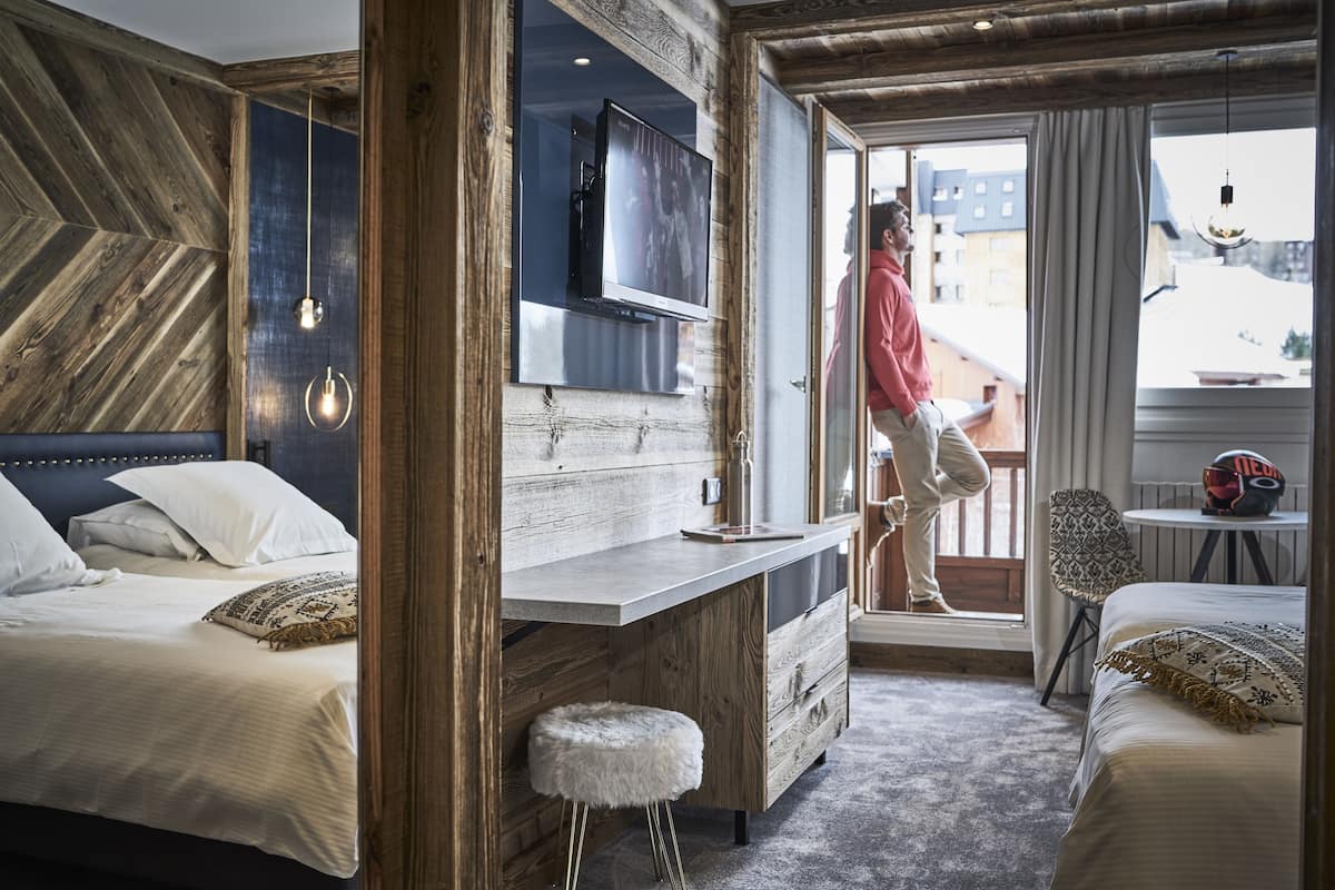 A hotel room with a wooden wall, a bed, a desk, a TV, and a balcony with a view.