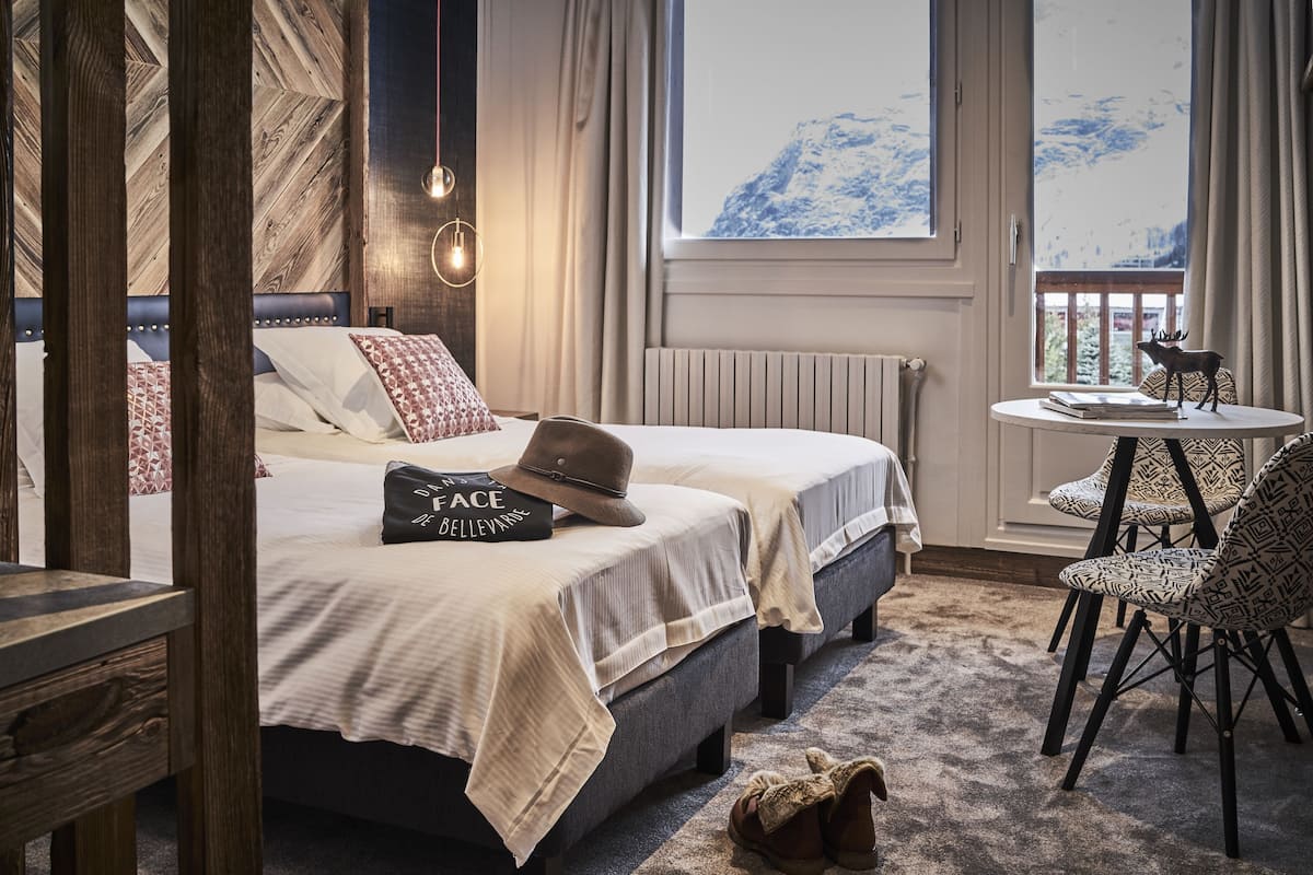 A bedroom with two beds, a view of mountains through the window, a small table with a book and a deer figurine, and a chair with a patterned design.