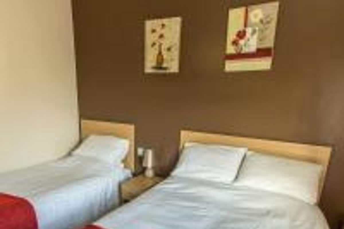 Une chambre d’hôtel avec deux lits simples, chacun agrémenté de linge de lit blanc et de touches de couleur rouge. On y trouve deux tables de chevet avec des lampes, et les murs sont décorés d’œuvres d’art encadrées.