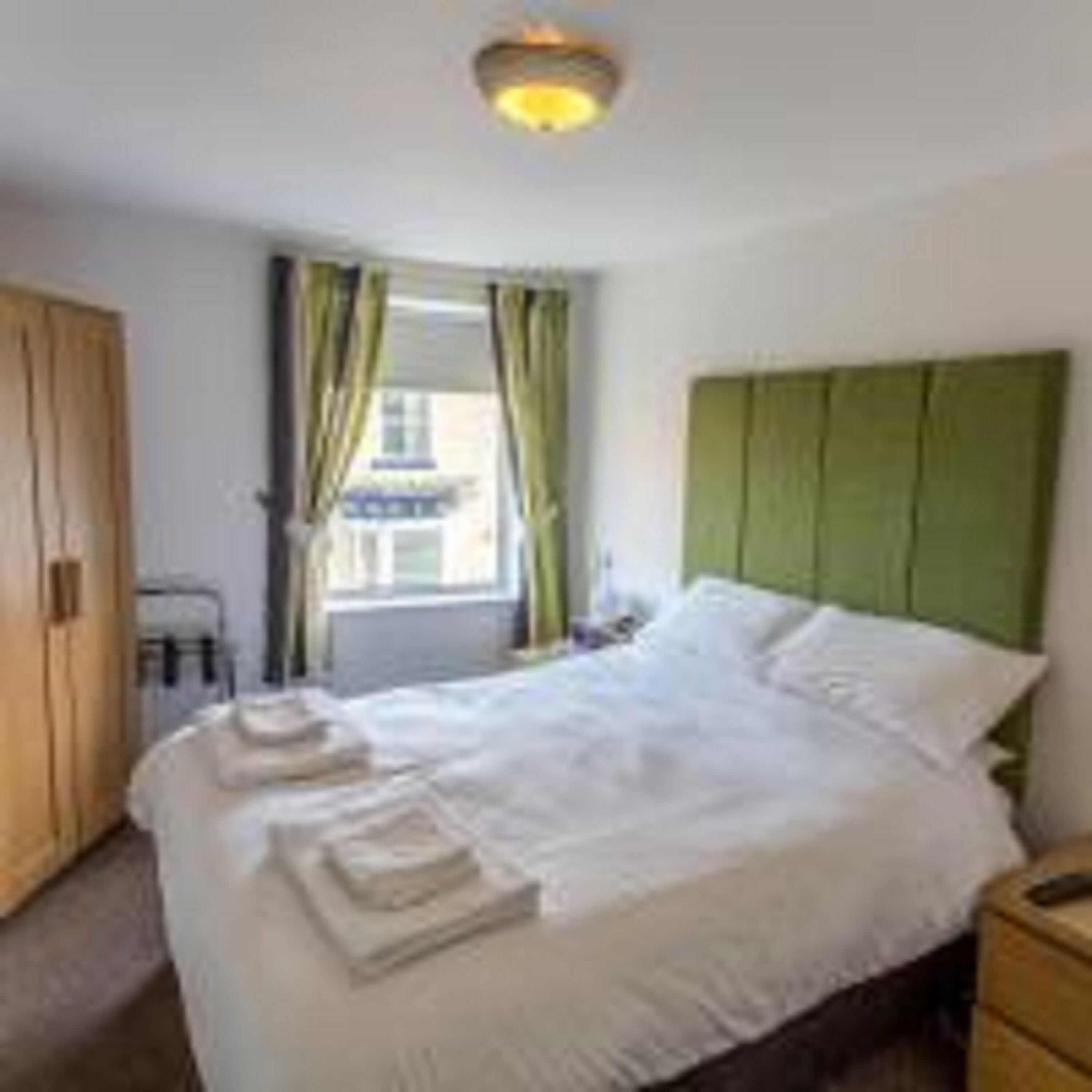A neatly made bed with white linens, a green headboard, and a window with curtains.