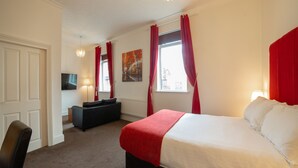 A hotel room with a large bed, a desk, a chair, a TV, and a window with red curtains. - The Douglas Hotel (Hartlepool)