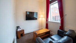 A living room with a flat-screen TV, a wooden coffee table, a sofa, a floor lamp, and a window with red curtains. - The Douglas Hotel (Hartlepool)