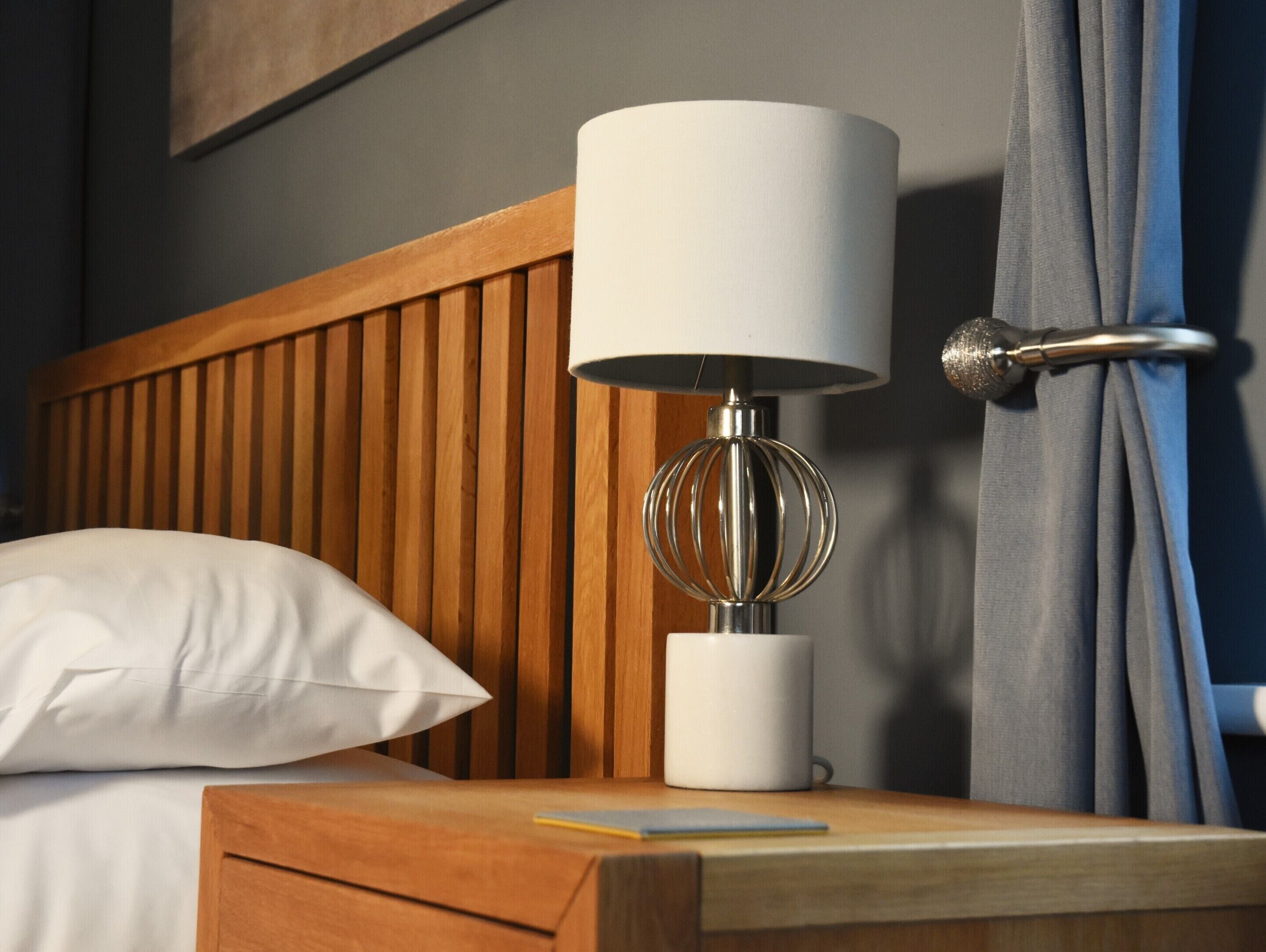 Un letto ben rifatto con una testiera in legno, un cuscino bianco, un comodino con una lampada e una tazza.