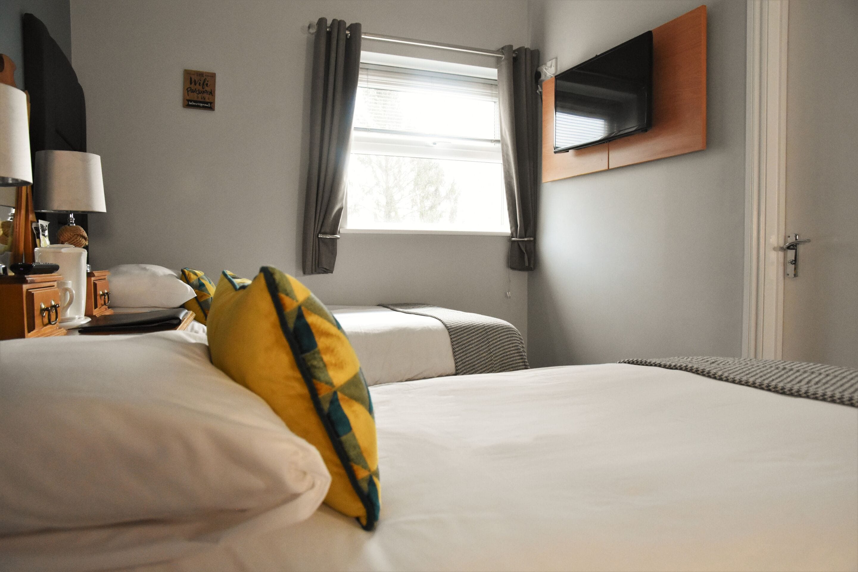 A hotel room with a bed, a bedside table with a lamp, a television mounted on the wall, and a window with curtains.