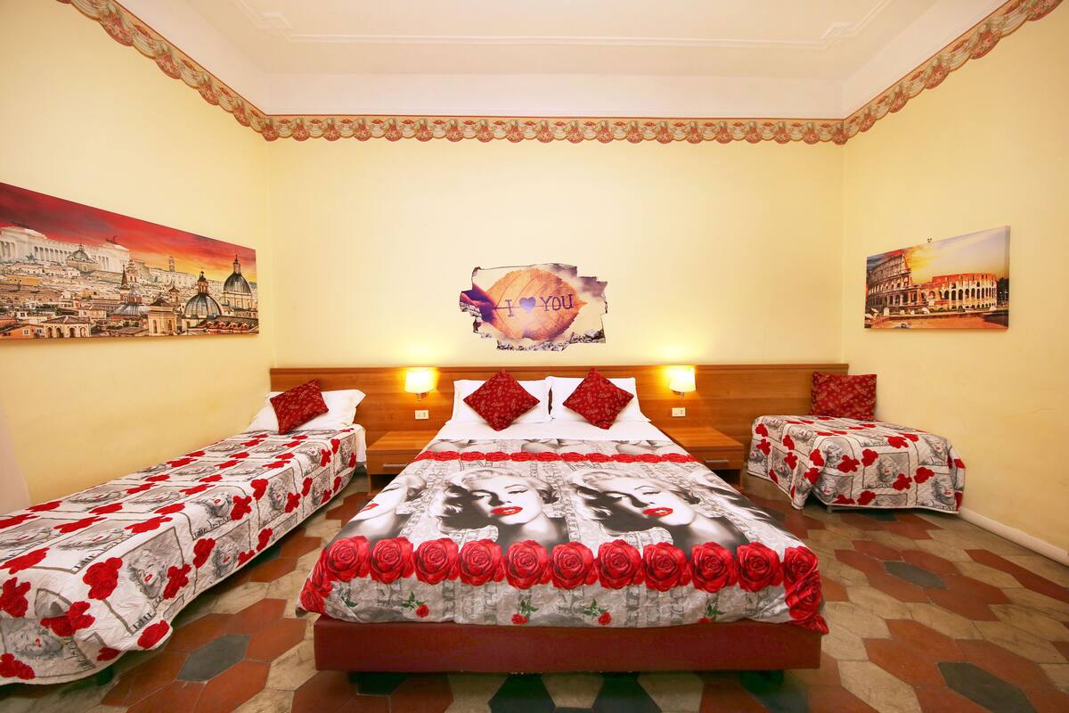 A hotel room with a large bed, two smaller beds, a headboard with a mirror, and a painting of a cityscape on the wall.