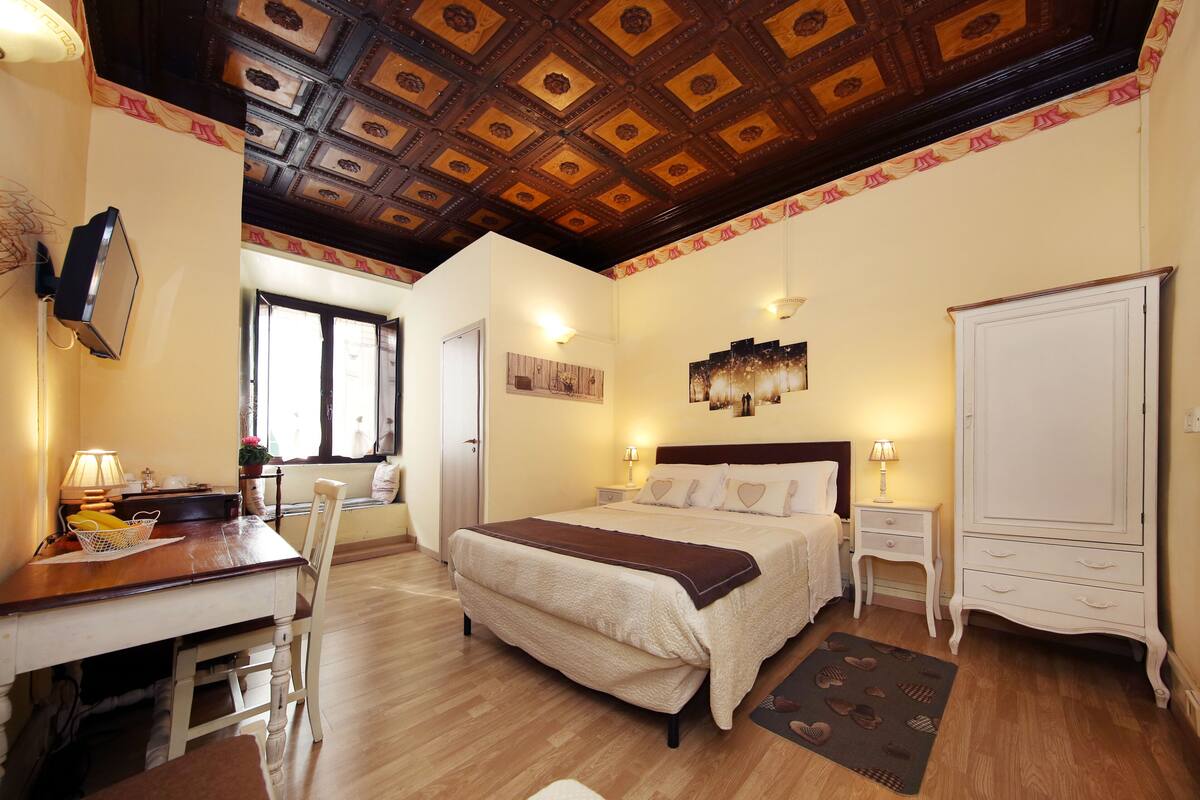 A hotel room with a wooden ceiling, a bed with a brown blanket, a desk with a lamp, a chair, and a wardrobe.