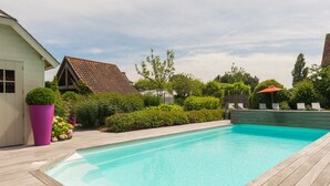 Una piscina con cubierta de madera, rodeada de follaje y una casa con tejado de tejas rojas.