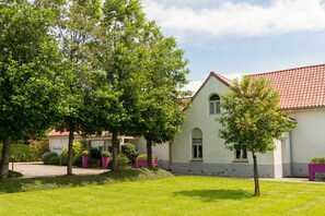 A white building with a red-tiled roof, surrounded by green trees and grass. - Duinhof (De Haan)
