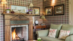 Una sala de estar acogedora con chimenea, papel tapiz escocés, un sofá verde con cojines florales y cuadros en la pared.