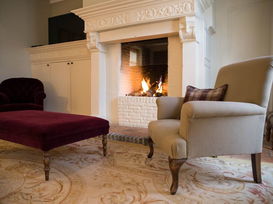 Una sala de estar con chimenea, un reposapiés rojo, un sillón color beige y una alfombra estampada.