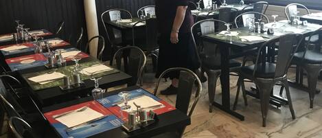 Interior de restaurante con mesas dispuestas para comer, sillas y una persona de pie cerca de las mesas.