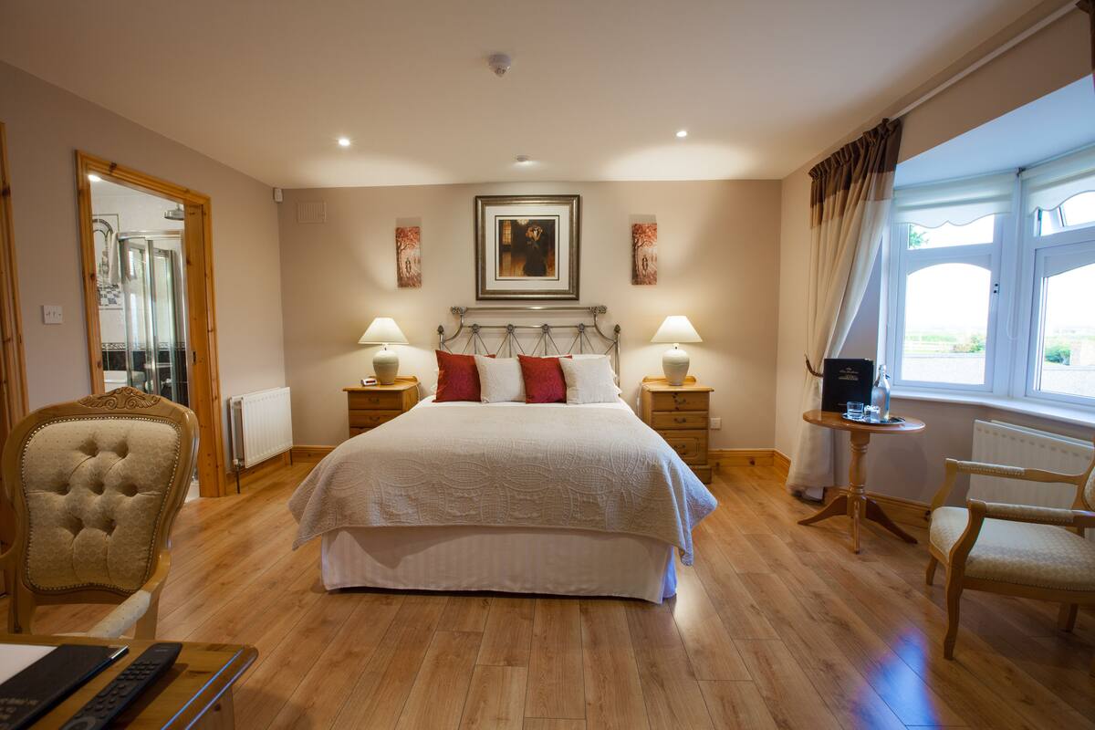 A spacious bedroom with a large bed, two bedside tables with lamps, a wooden chair, and a view of the outdoors through a window.