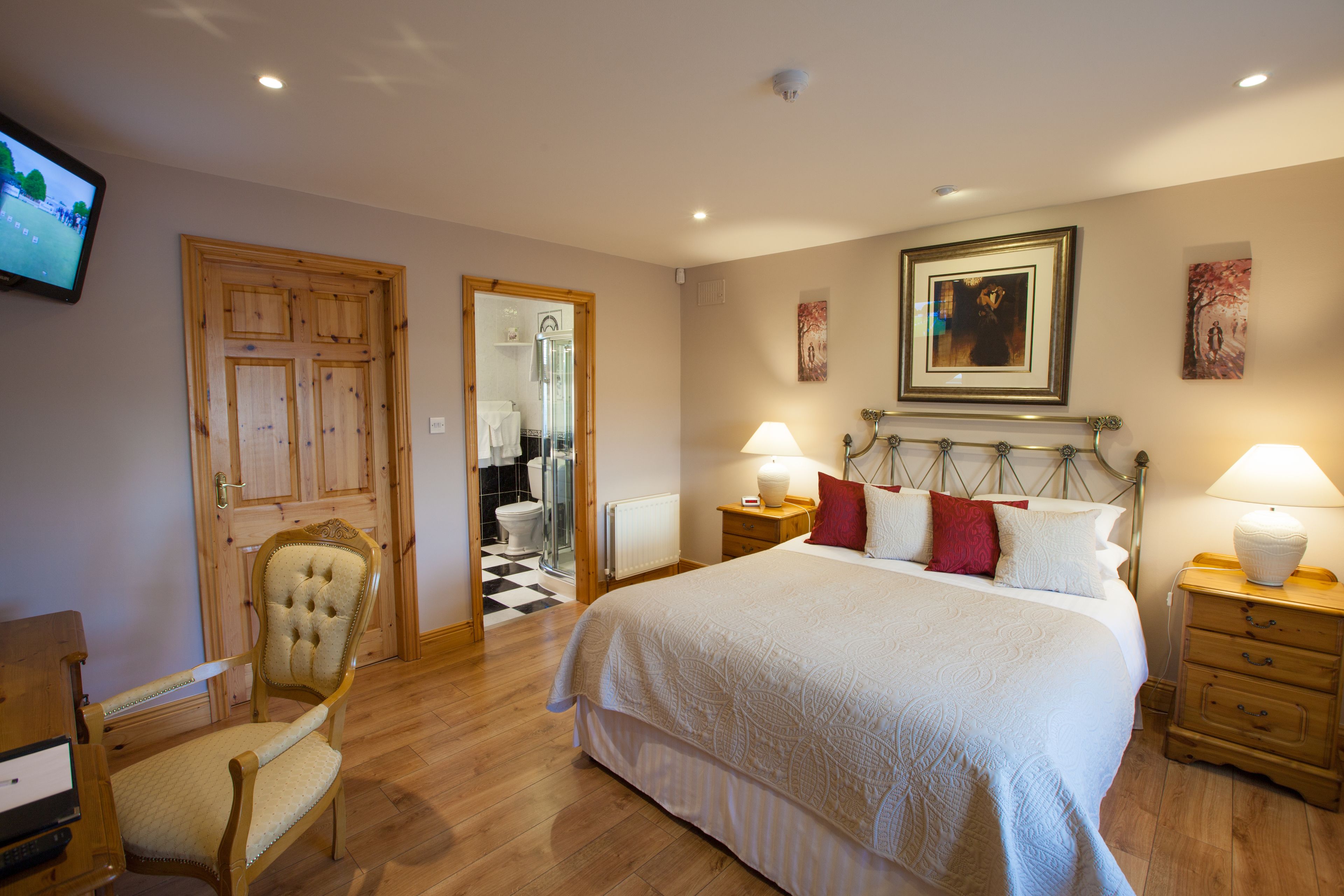 A bedroom with a large bed, wooden floor, a chair, a television, and a bathroom visible through an open door.