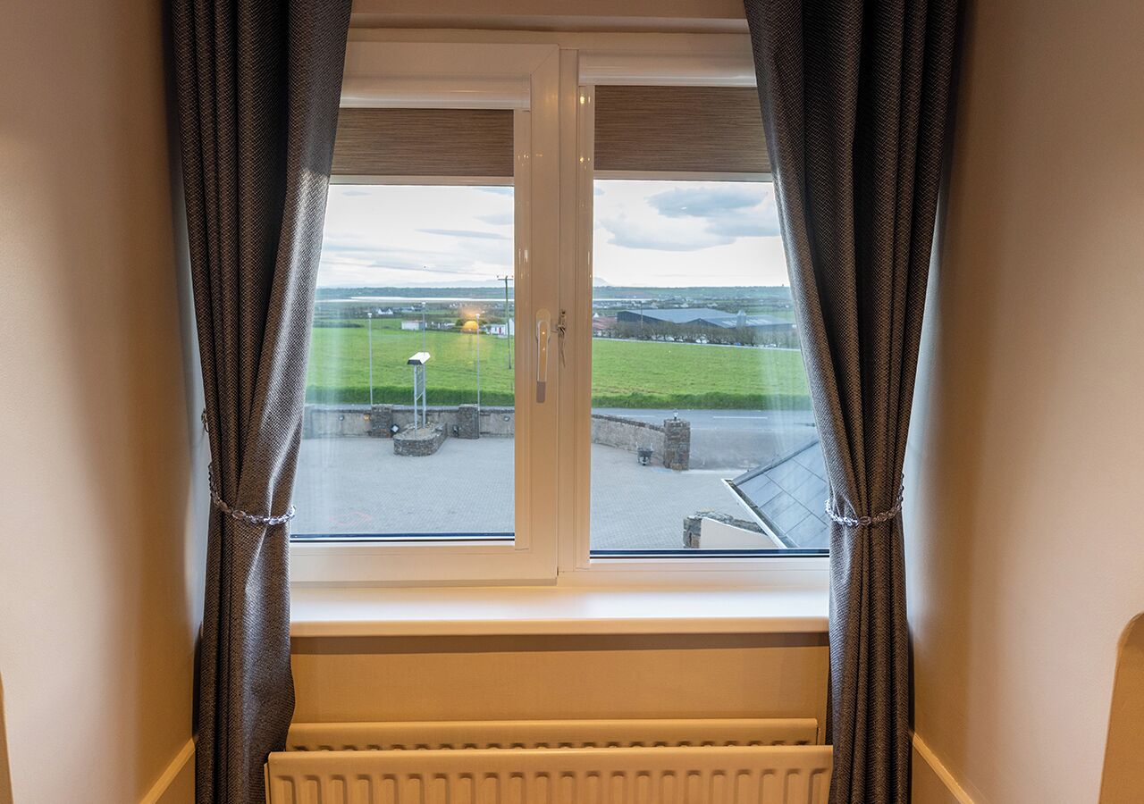 Una ventana con cortinas, un radiador en la parte inferior y vista a un paisaje verde con edificios.