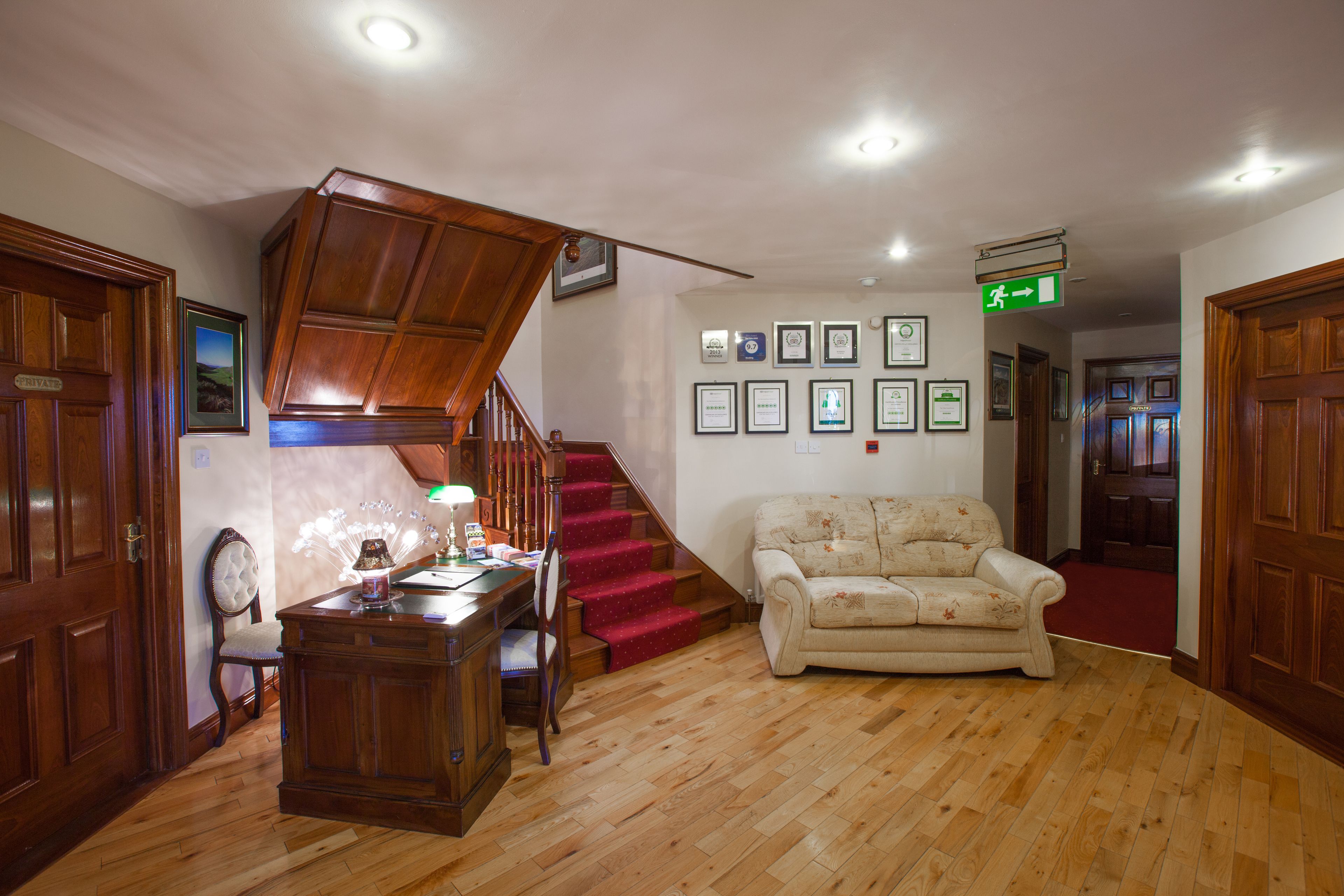 A spacious living area with a wooden staircase, a sofa, a desk with a lamp, and framed certificates on the wall.