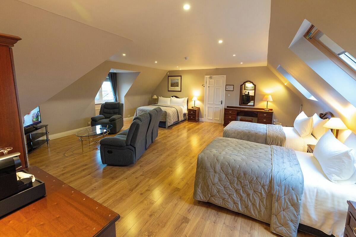 A spacious room with a sloped ceiling, two beds, a seating area with a sofa and chairs, a television, and a wooden floor.