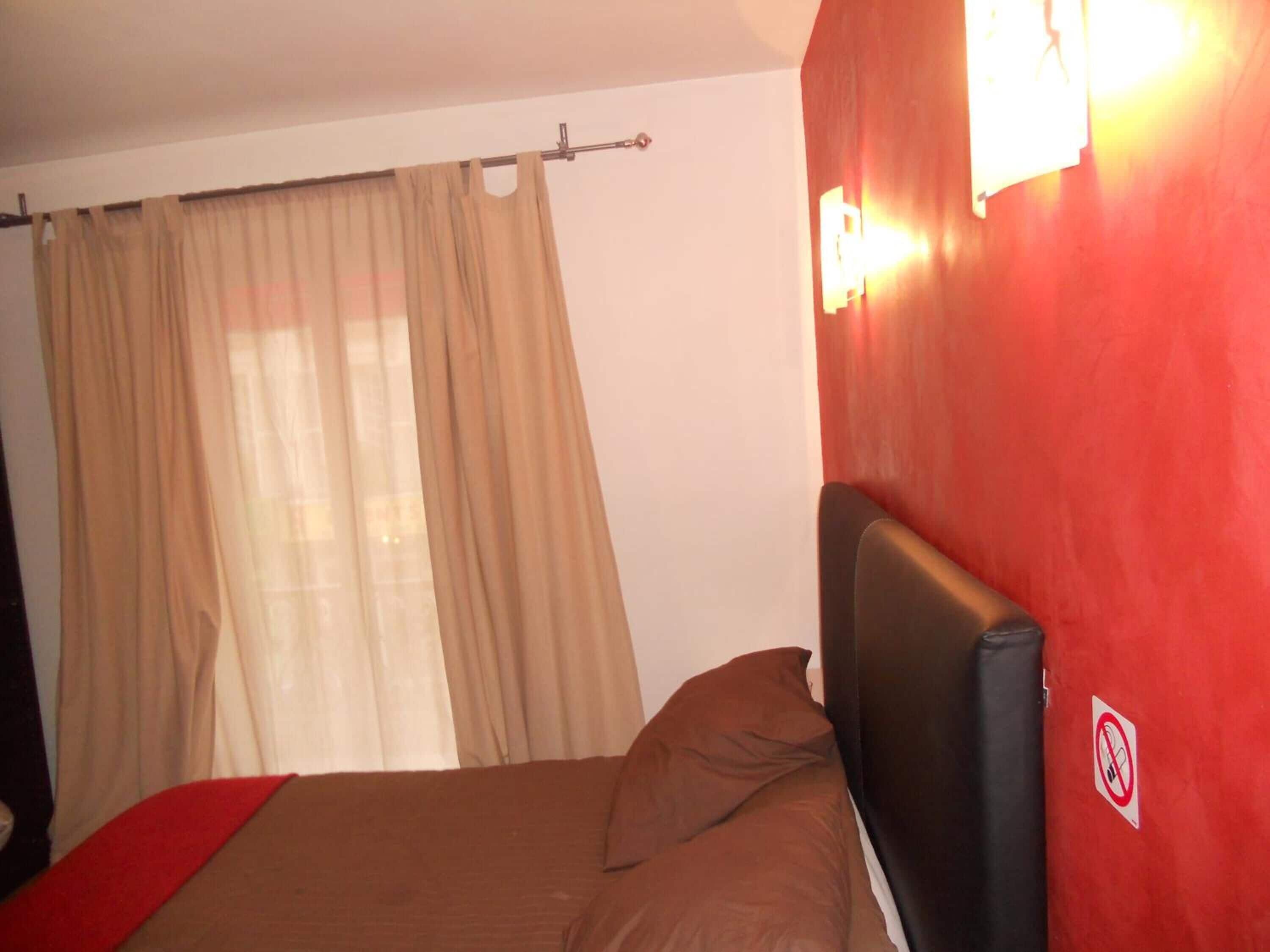Une chambre à coucher avec un lit, des rideaux beiges, un mur rouge et un panneau indiquant qu’il est interdit de fumer.