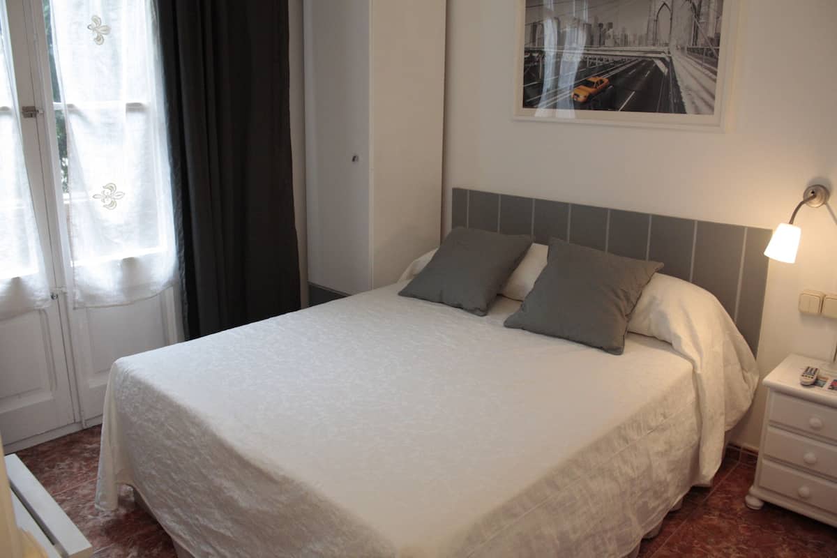 Una cama bien hecha con un edredón blanco y dos almohadas, una mesita de noche con una lámpara, y una ventana con cortinas.