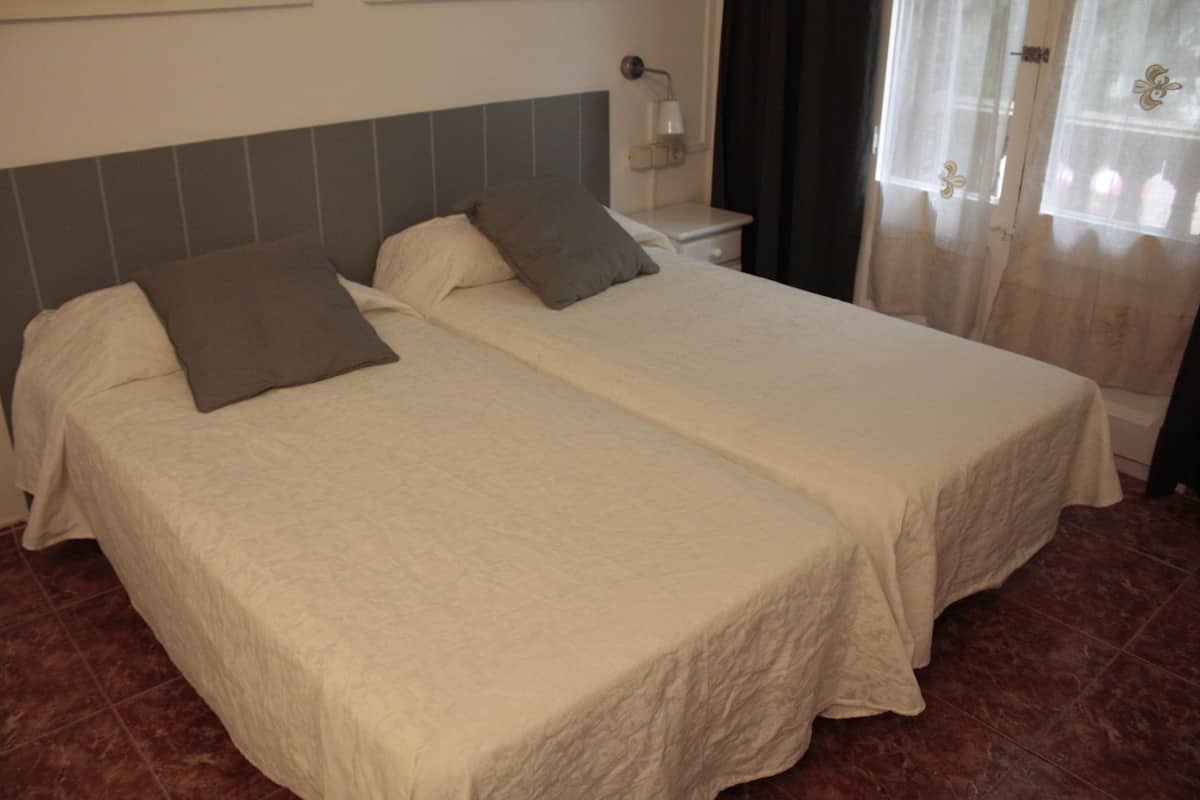 Una cama doble con dos almohadas, una mesita de noche con lámpara y una ventana con cortinas.